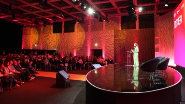 Eine Frau im grünen Hosenanzug steht auf einer runden Bühne und spricht vor einem Publikum bei einer Konferenz. 