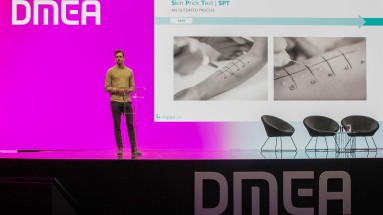 Eine Person steht auf einer Bühne. Neben ihm eine Präsentation mit Fotos eines Allergietests am Arm. 